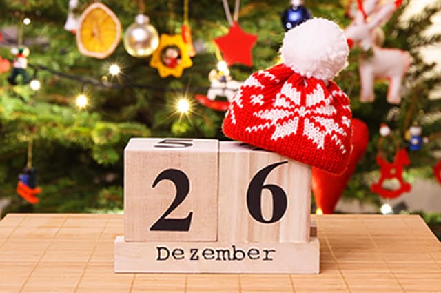 Stefanitag: Datum auf Holzwürfel mit einer roten weihnachtlichen Wollmütze oben drauf und einem geschmückten Weihnachtsbaum im Hintergrund mit Lichtern